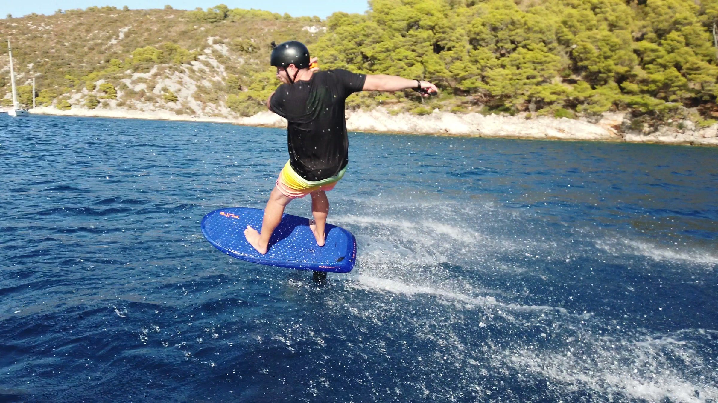 A rider in a helmet flies an eFoil close to the camera, arms out for balance, in a sunny bay with pine trees.