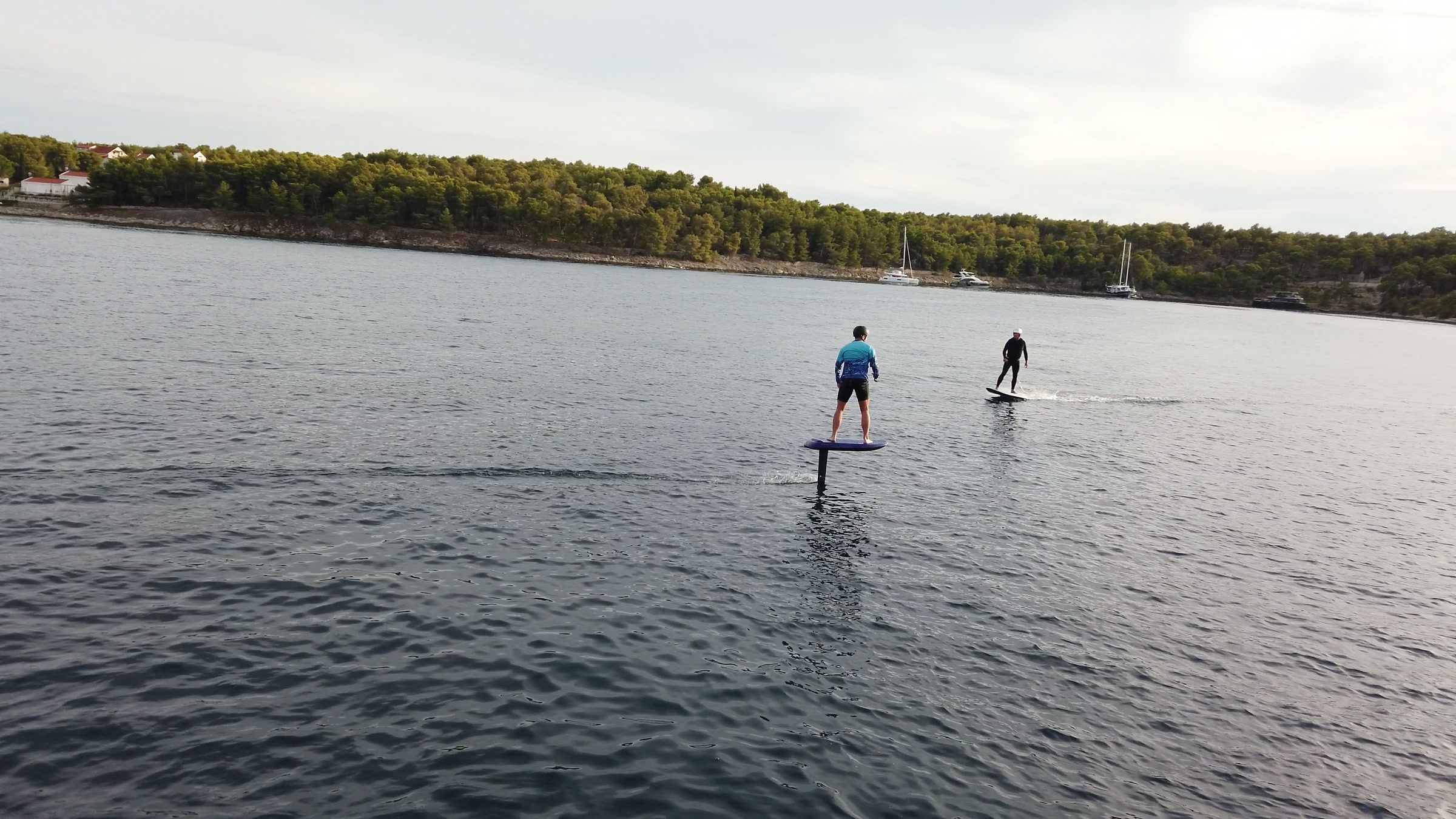Two riders fly on eFoils side by side across a calm bay lined with pine trees and anchored sailing boats.