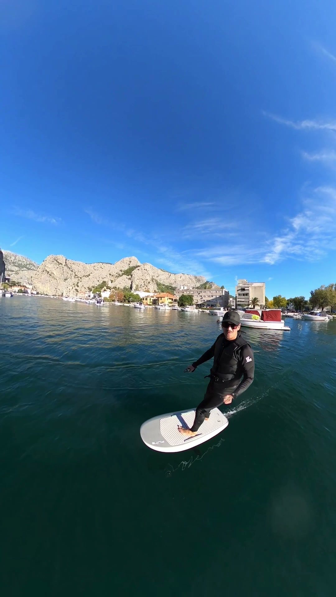 A smiling rider carves an eFoil through the green water of Omiš harbour, with moored boats, the town and limestone cliffs behind.