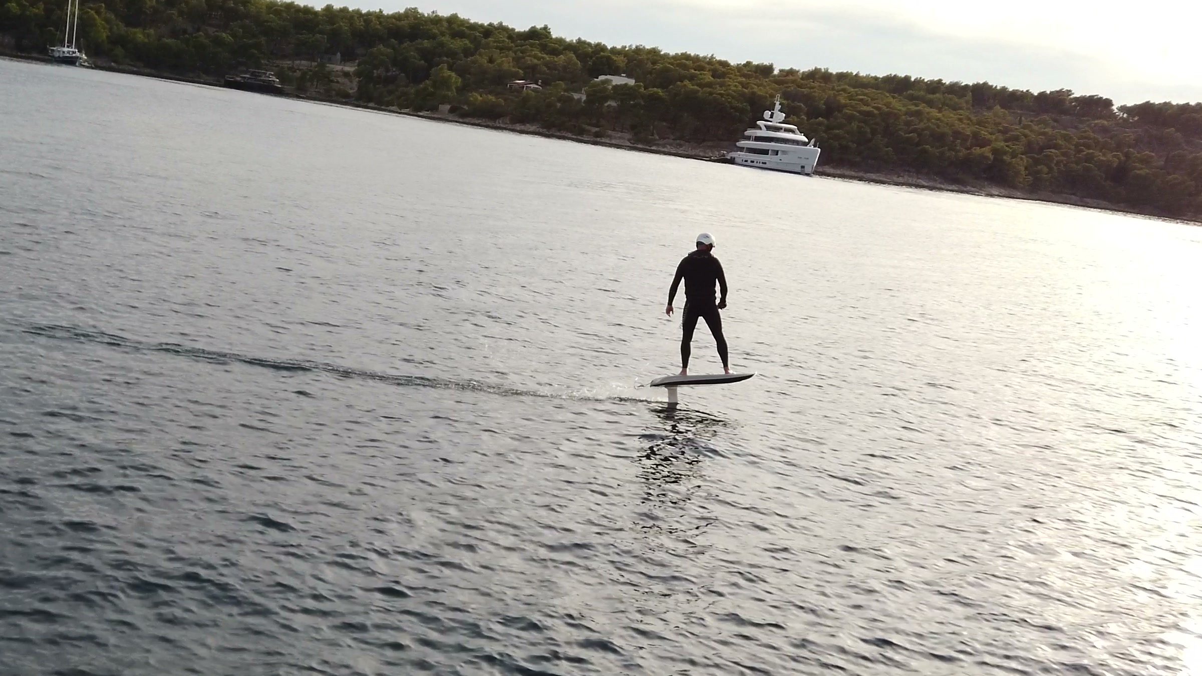 A rider flies an eFoil across sunlit water towards a large yacht anchored off a wooded shore.