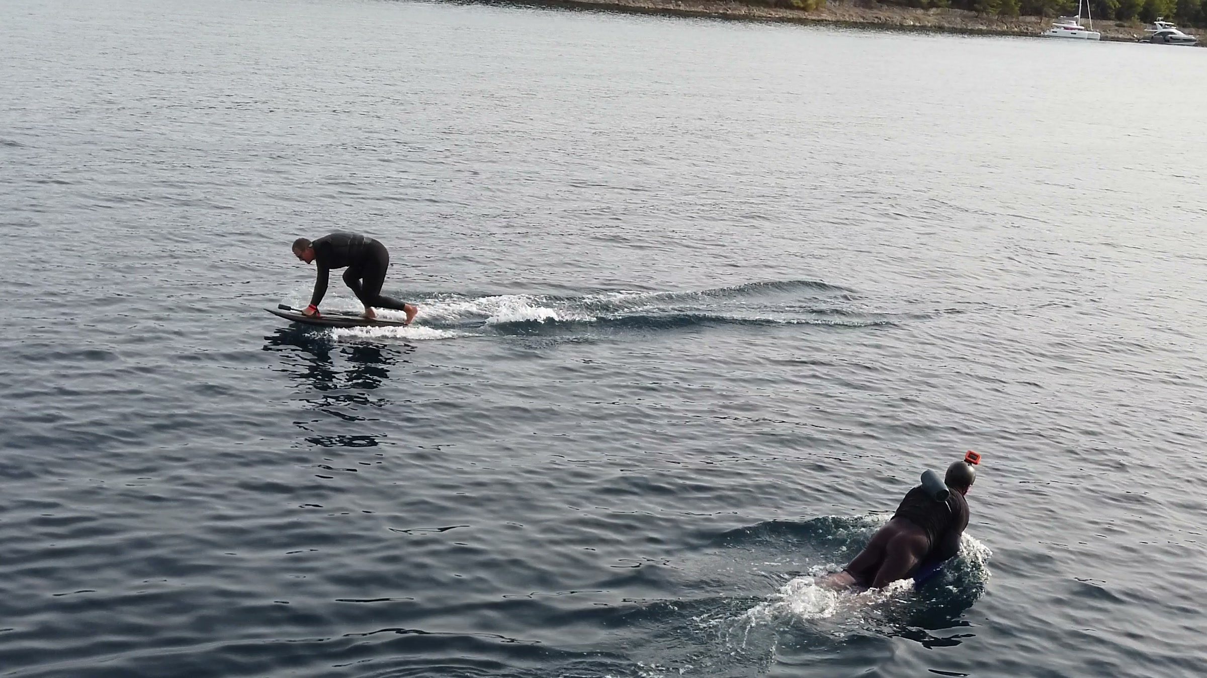 A beginner rises to his knees on an eFoil board in a calm bay while another guest paddles out lying on his board.