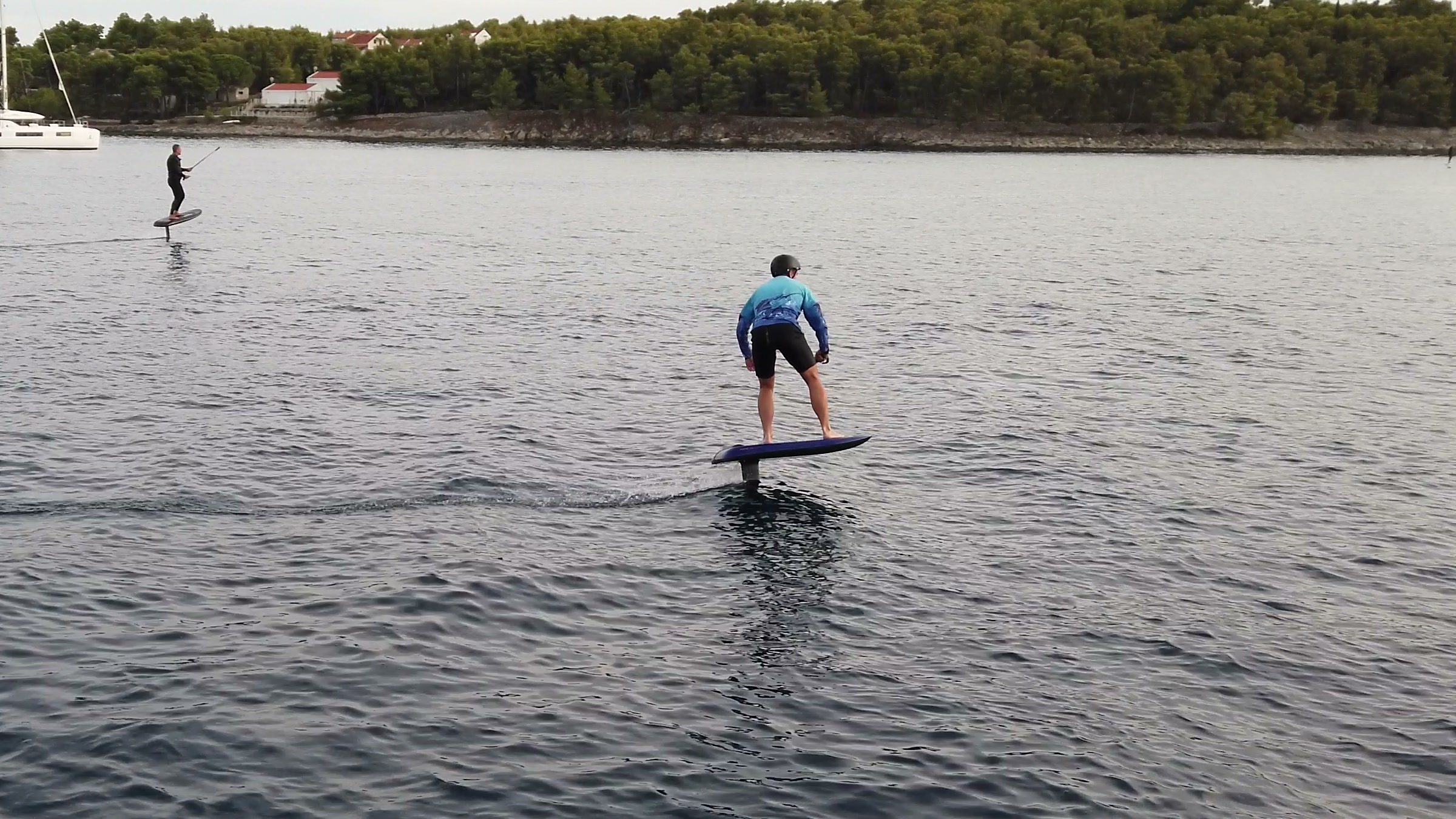 A guest in a blue top flies an eFoil steadily across a calm bay while the instructor rides further out.