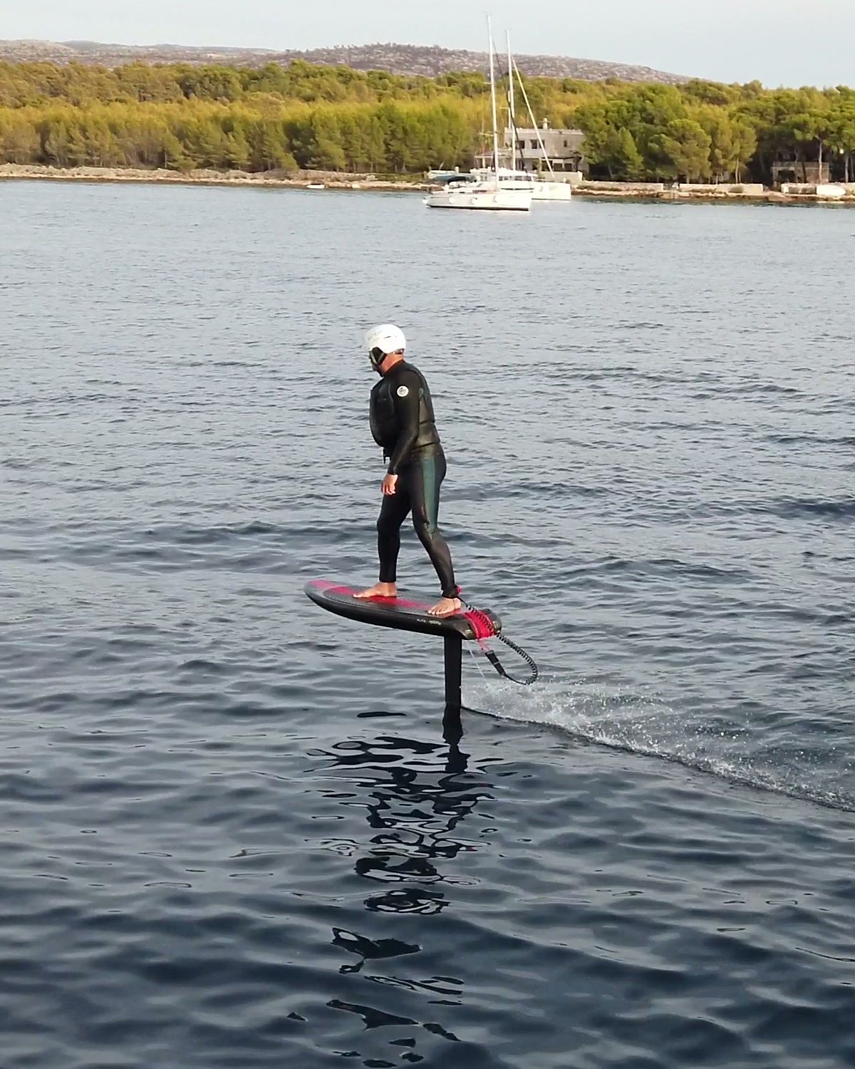 A rider in a white helmet flies a red eFoil board above calm water, the foil mast rising clear of the surface, with sailboats behind.