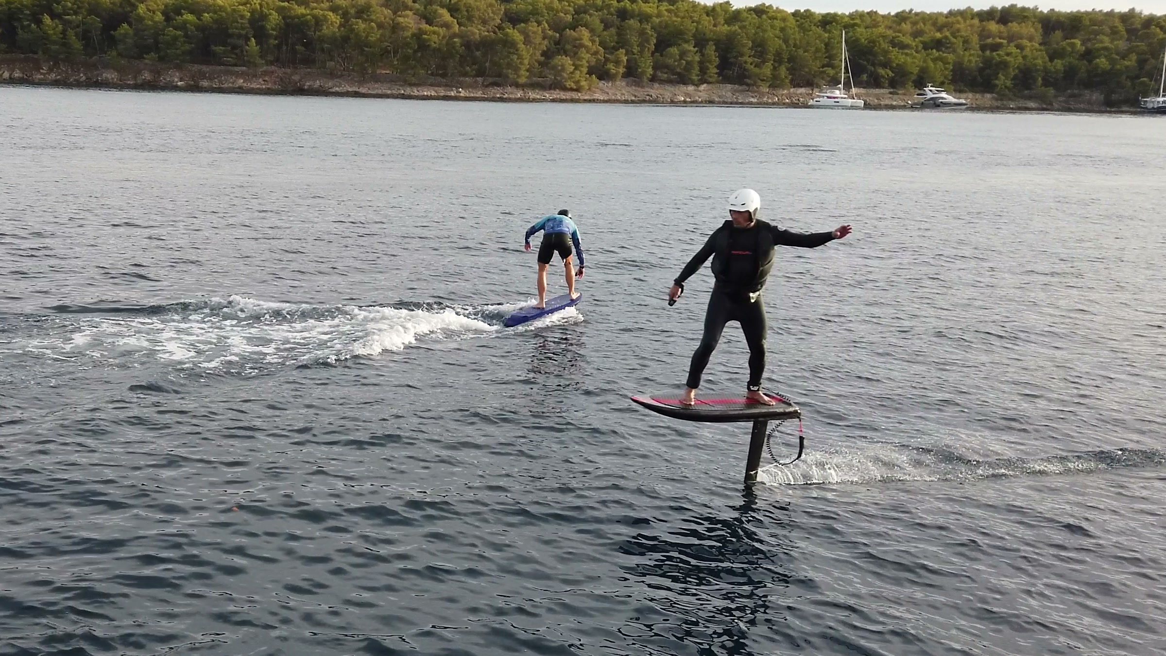 An instructor and a guest fly their eFoils side by side across a calm bay lined with pine trees.