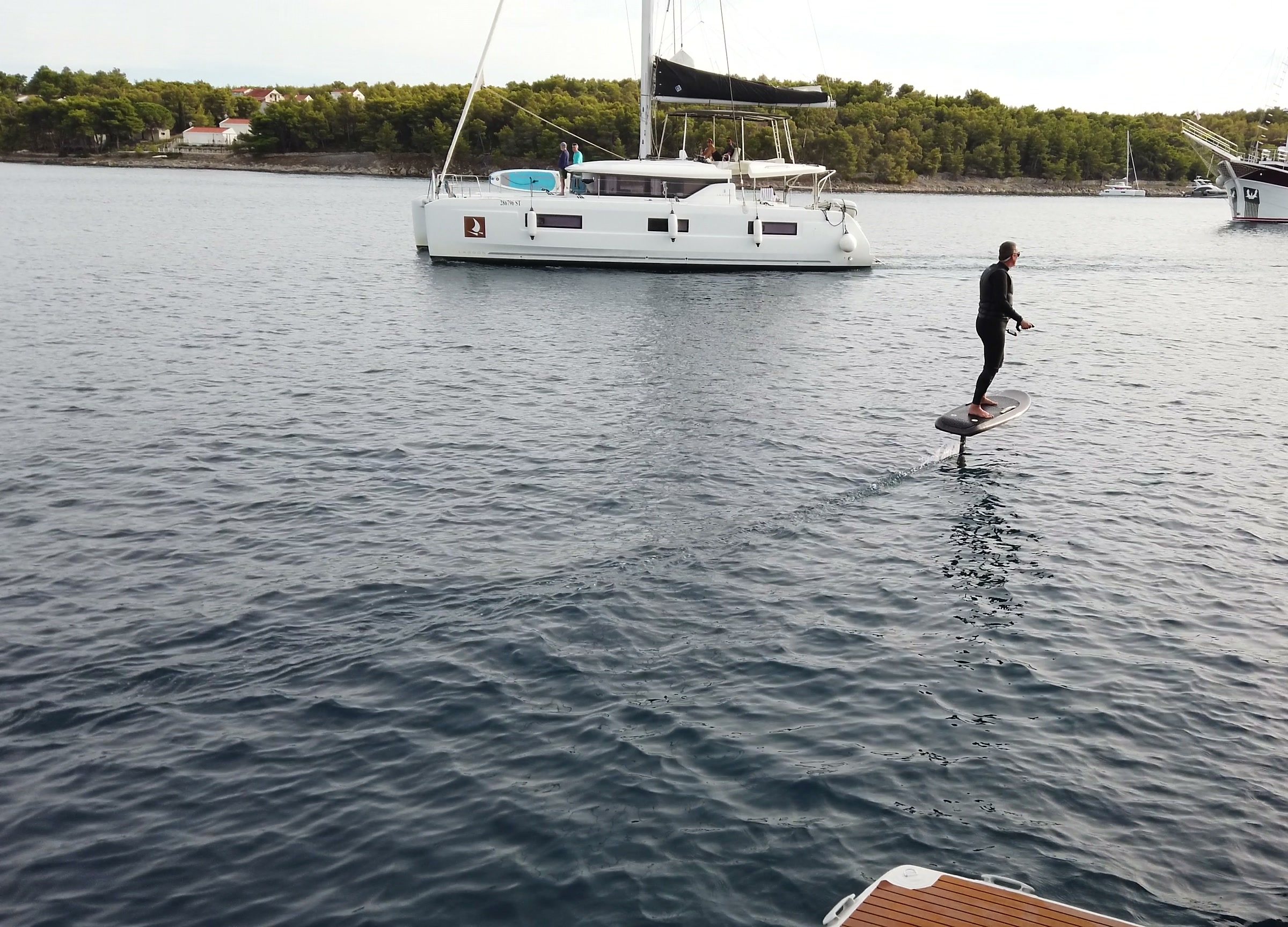 A rider flies an eFoil past a catamaran and a gulet anchored in a green bay.