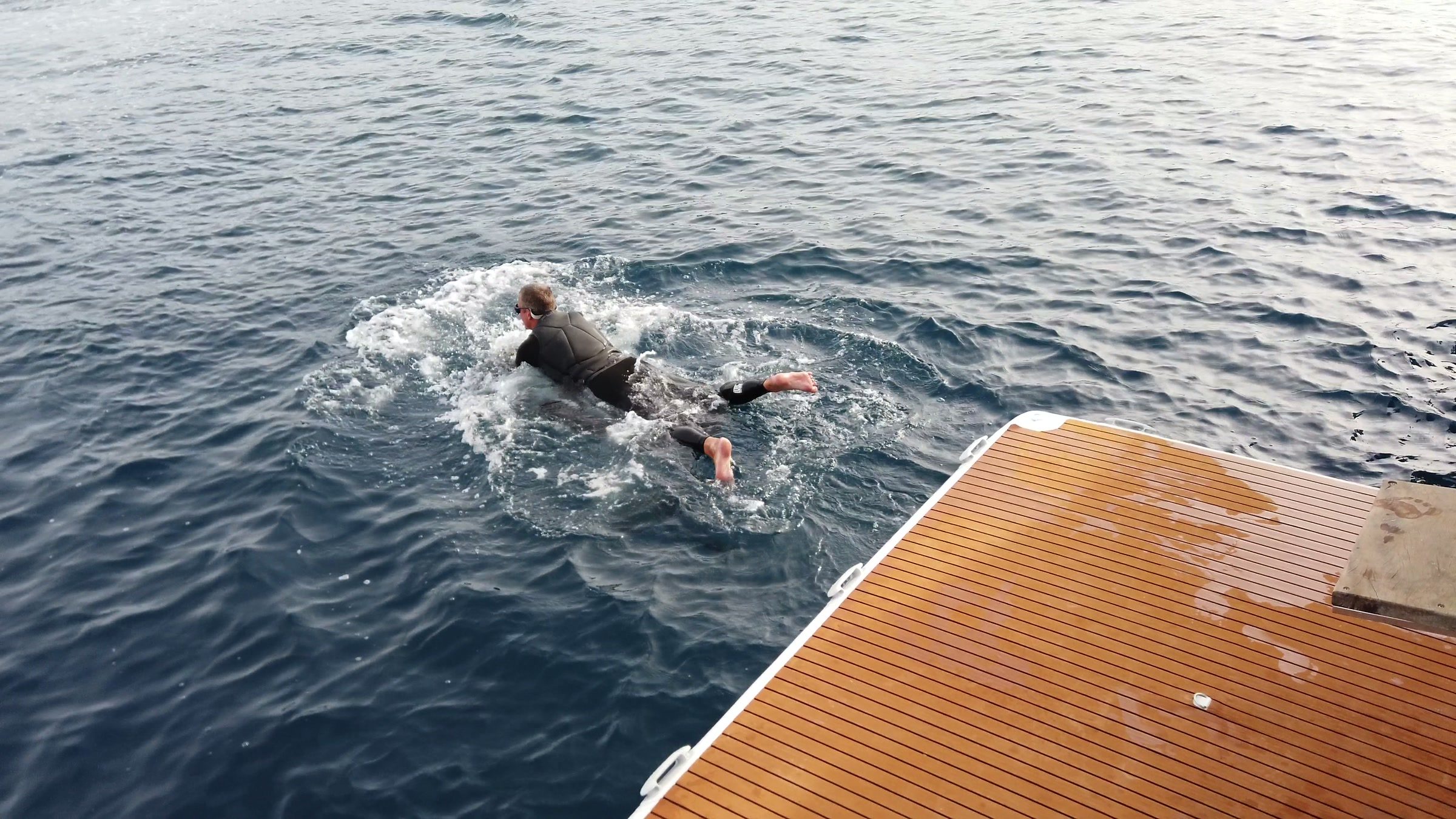 A guest lying on an eFoil board paddles away from the teak swim platform of a boat.