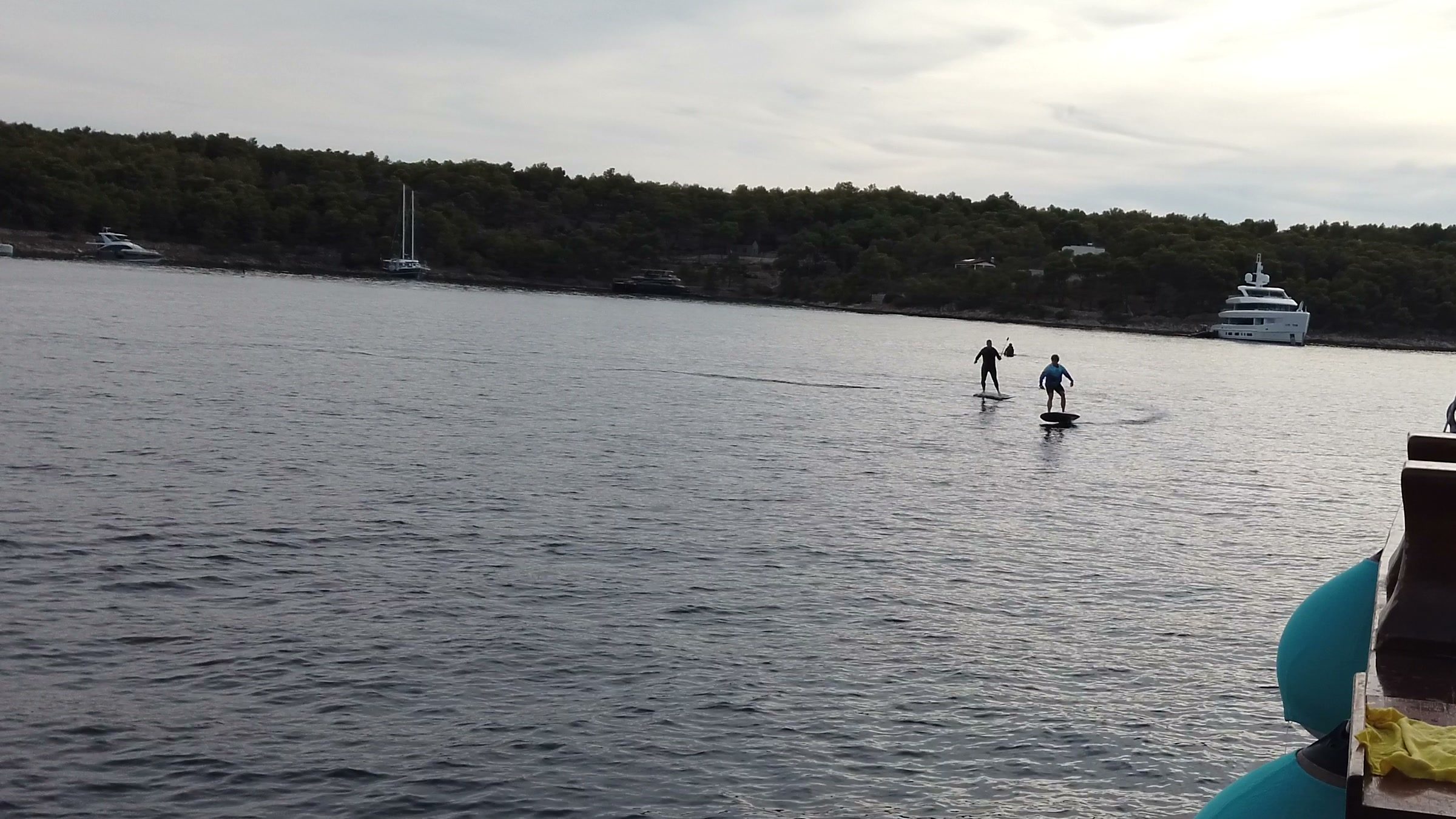 Two eFoil riders fly side by side across a quiet bay lined with pine trees, a yacht anchored behind them.