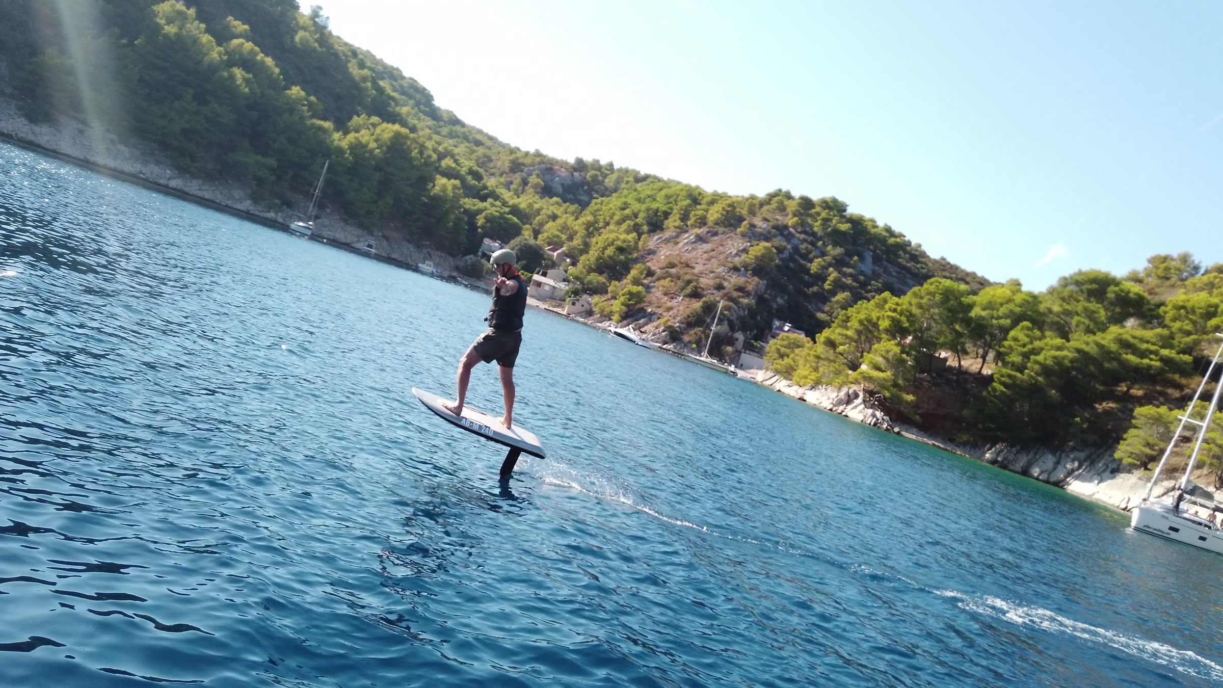 A rider flies an eFoil in a turquoise bay beside a pine-covered shore, sailing yachts at anchor.