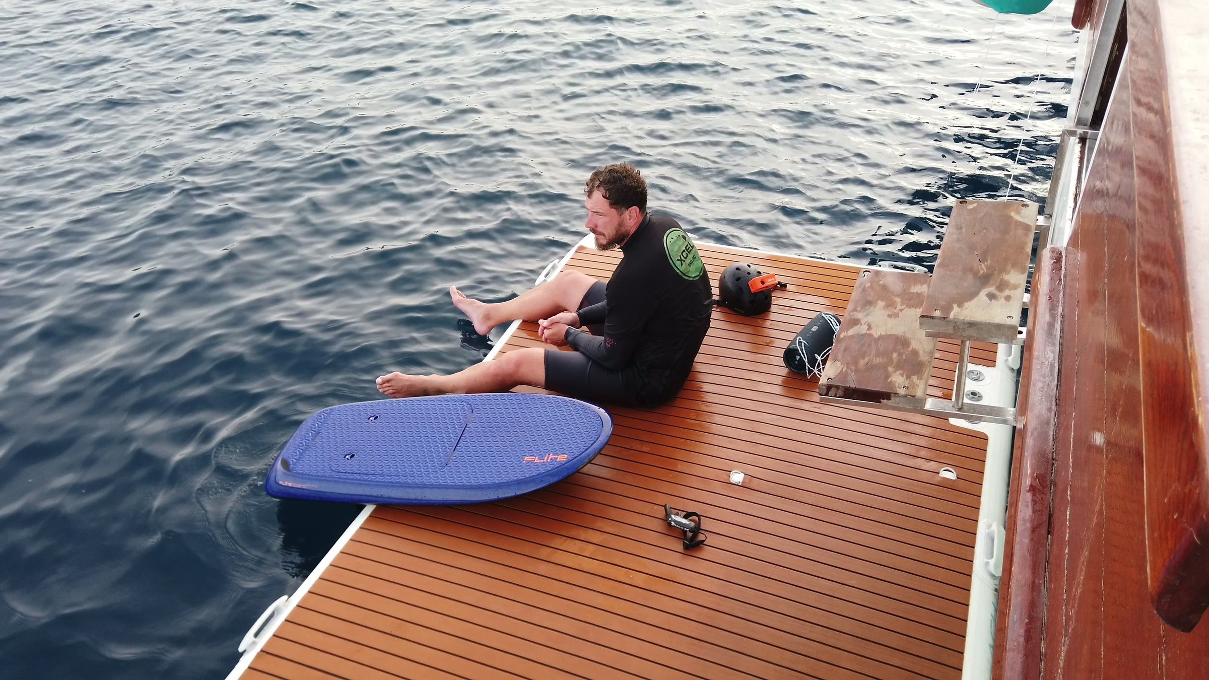 A guest sits on a boat's teak swim platform next to a blue eFoil board, ready to ride.