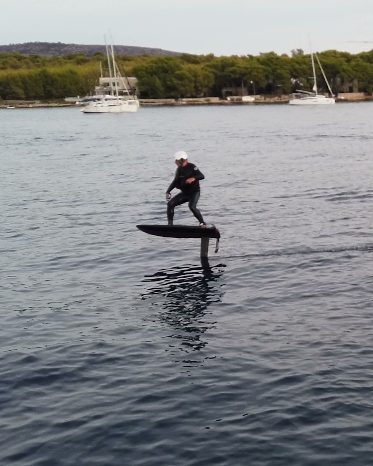 A rider leans into a turn on a black eFoil board flying above a calm bay, the mast visible beneath it.
