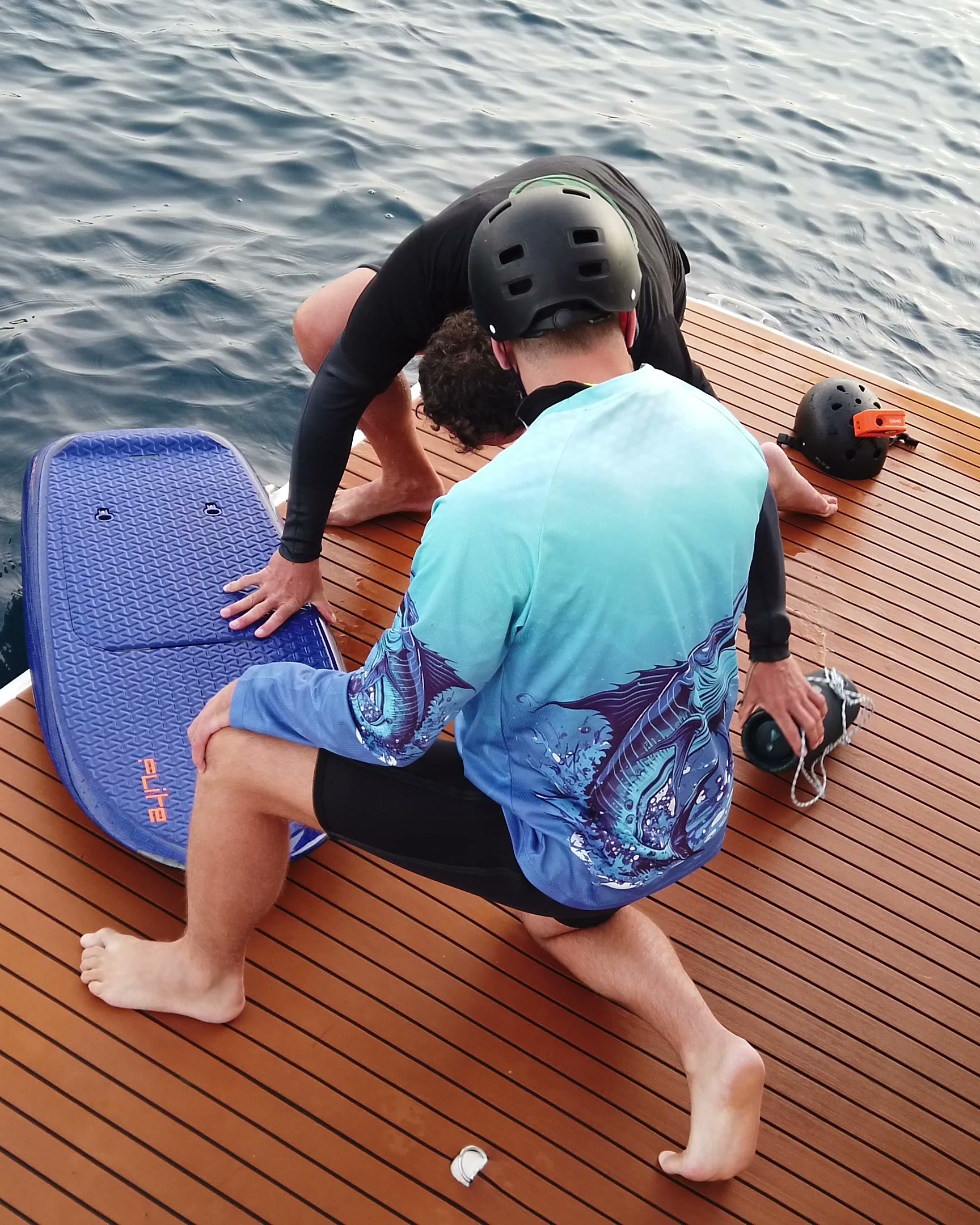 On the swim platform of a boat, an instructor helps a guest in a helmet get ready with a blue eFoil board.