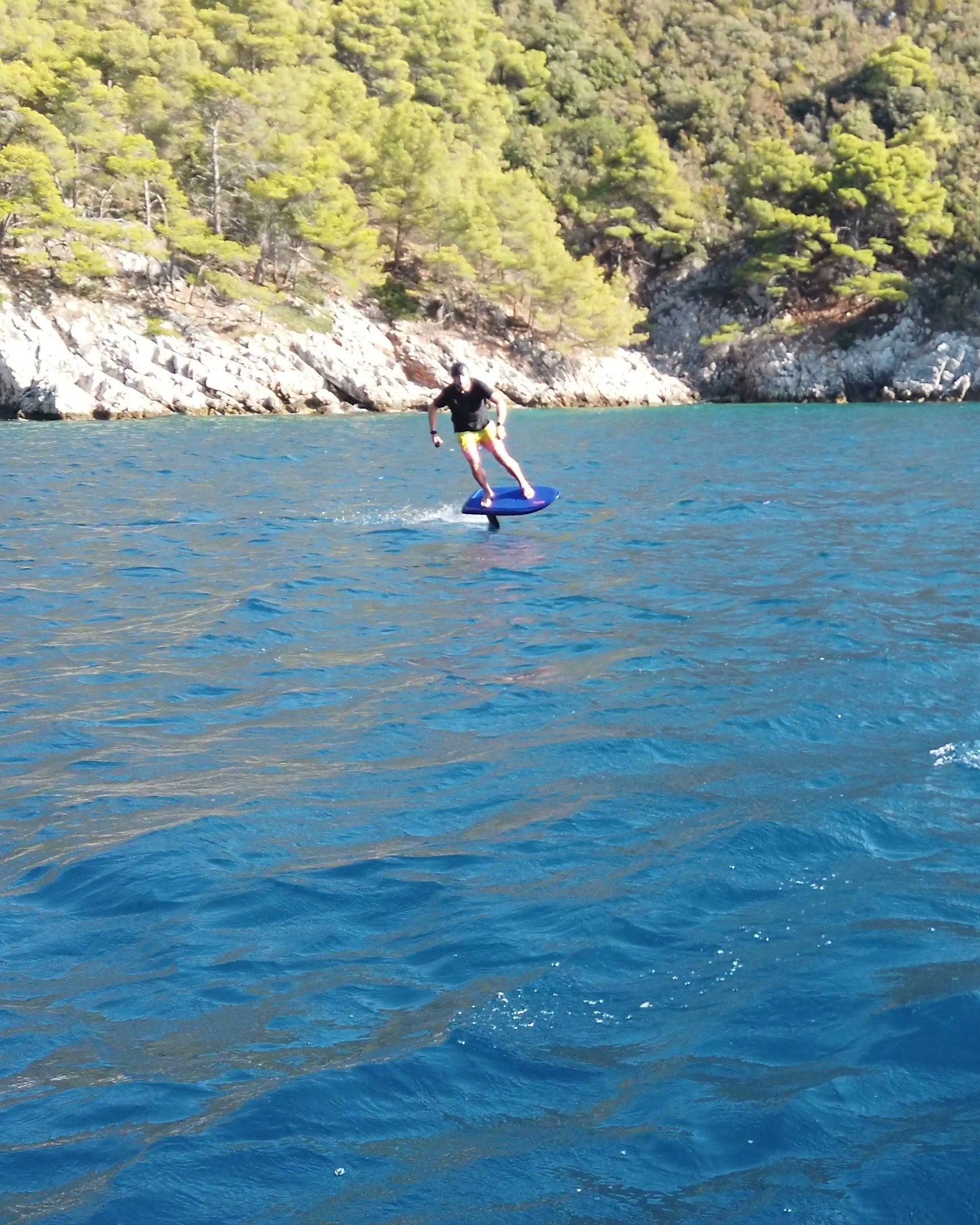 A rider turns an eFoil over clear turquoise water along a pine-covered, rocky shore.