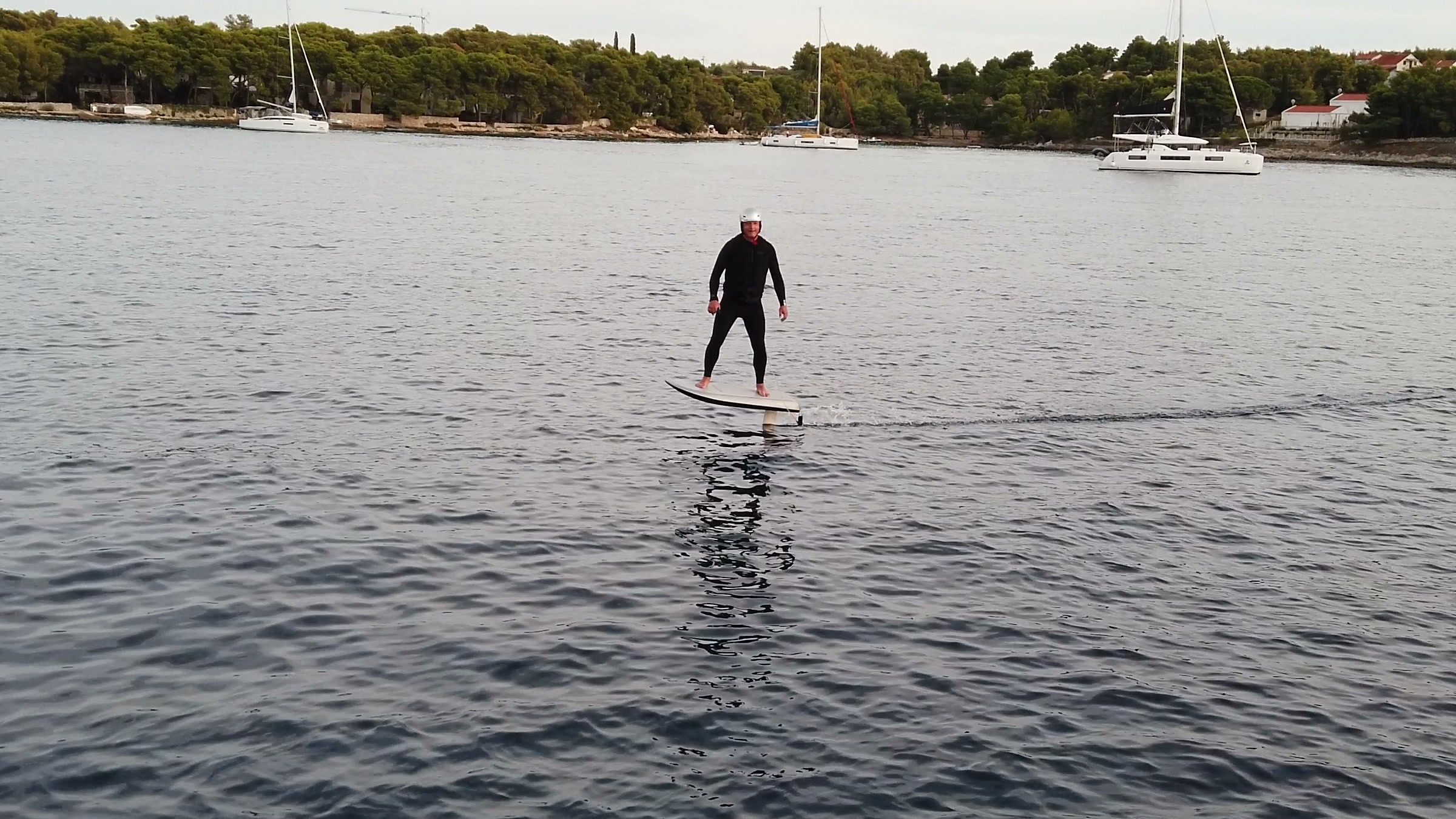 A rider stands tall on an eFoil flying over glassy water, reflected below, with sailboats at anchor behind.