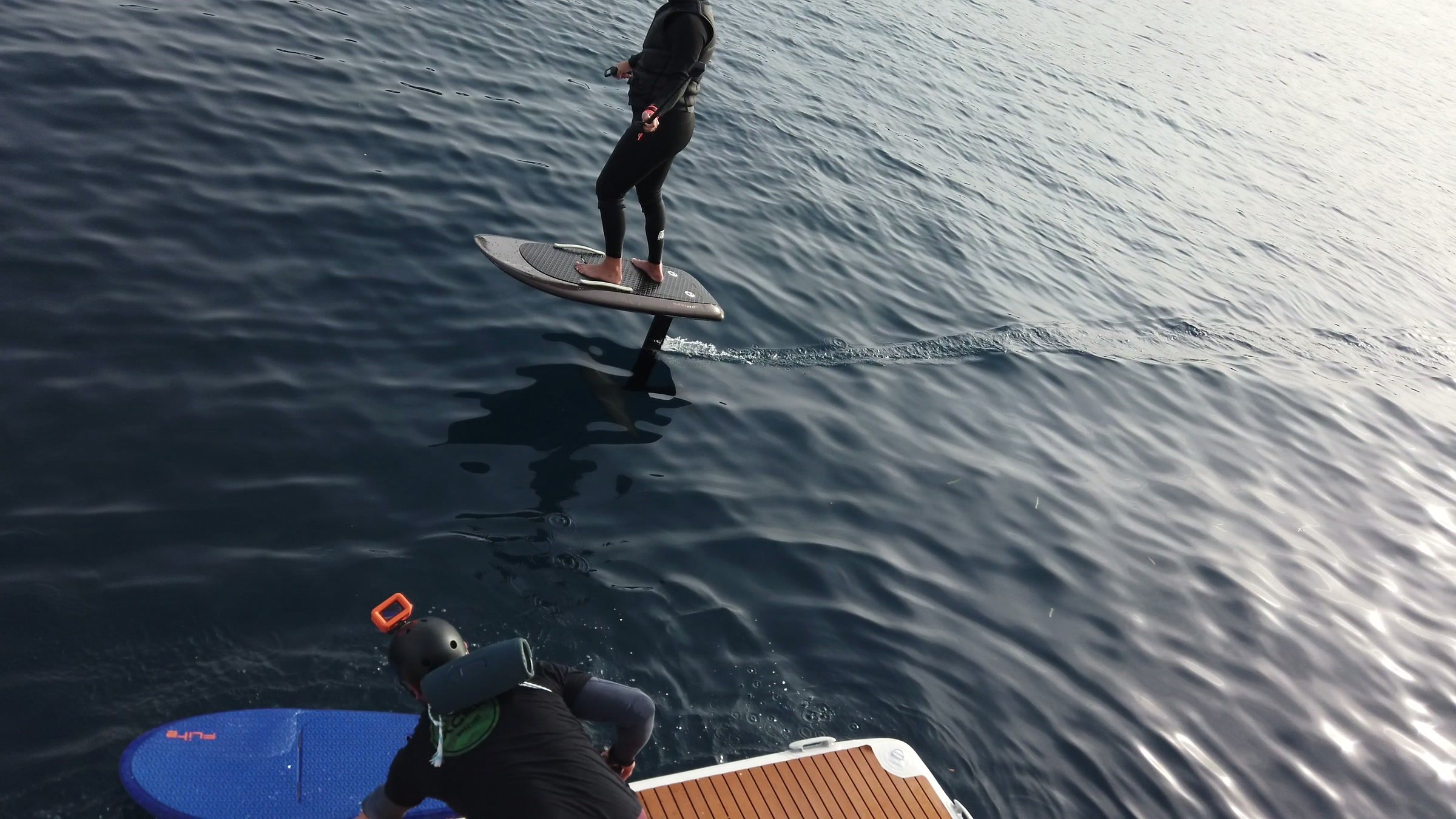 A rider flies an eFoil past the swim platform of a boat, where the next guest waits with a board for their turn.