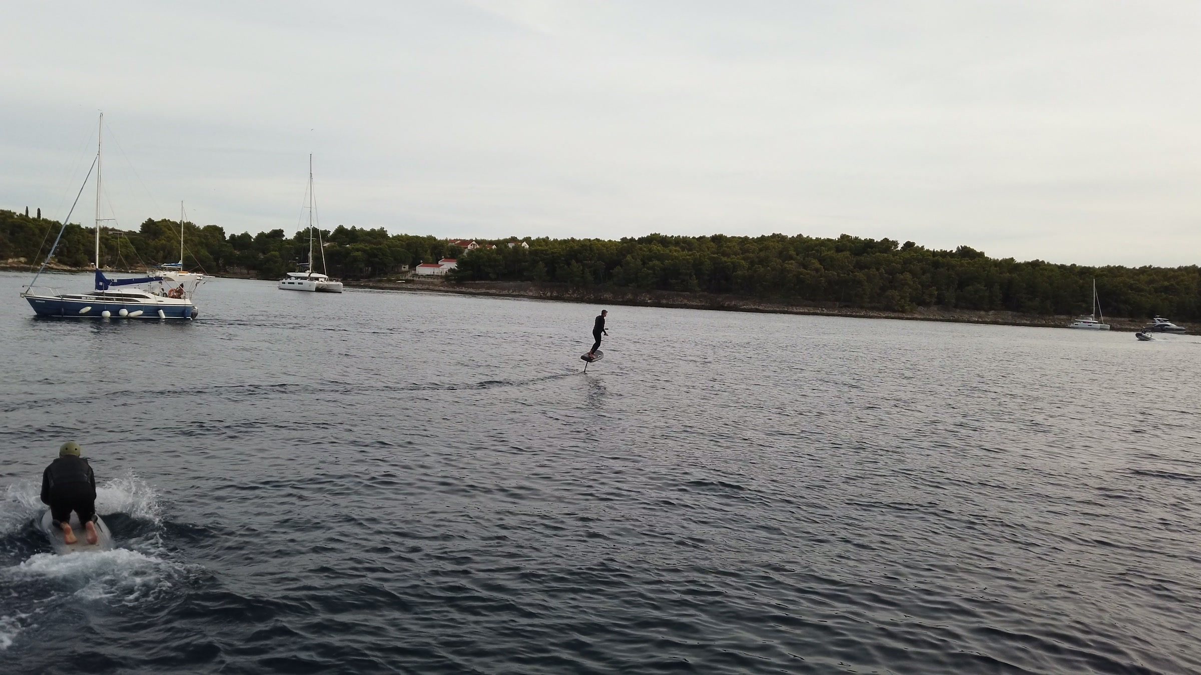 A rider flies an eFoil past anchored sailboats while a guest behind him gets up on his board.