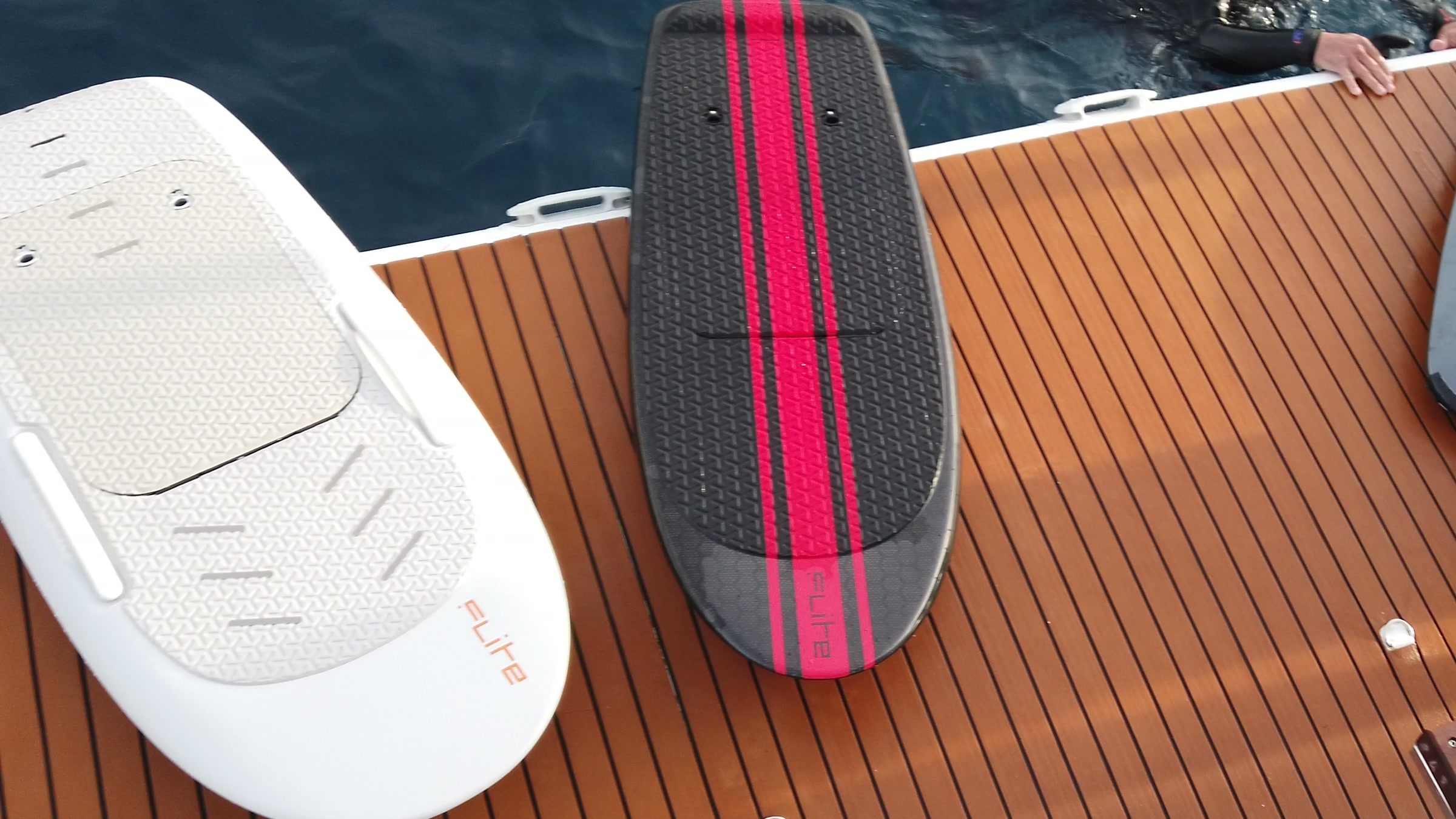 Two eFoil boards, one white and one black with red stripes, lie on a teak boat deck beside the sea.