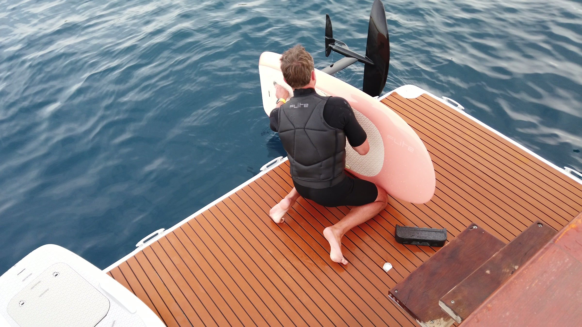 On a boat's swim platform, a rider in a wetsuit gets an eFoil board ready, the foil wing raised beside him.