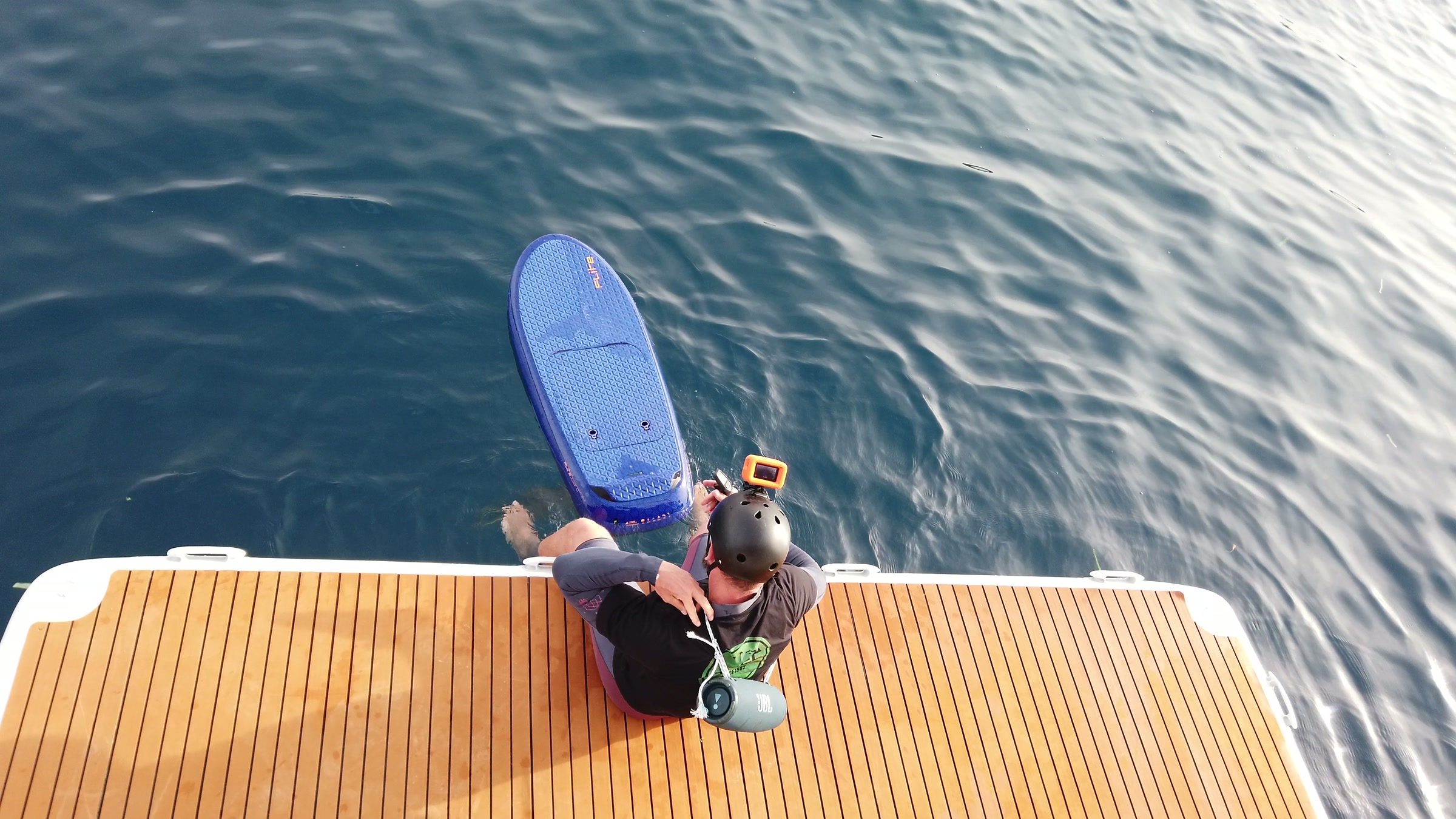 Seen from above, a rider in a helmet sits at the edge of the swim platform with the board in the water.