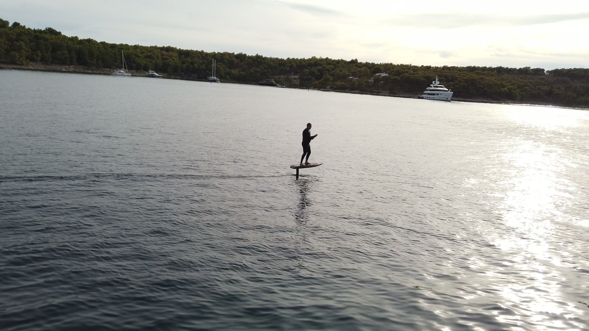 A rider stands on an eFoil and glides across a calm bay towards the low sun glittering on the water.