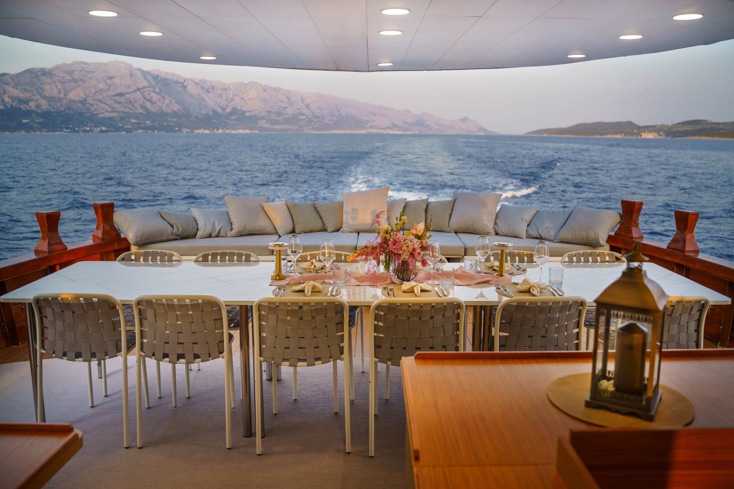 A long table laid on the aft deck, cushioned seating behind it and the wake stretching towards the mountains.