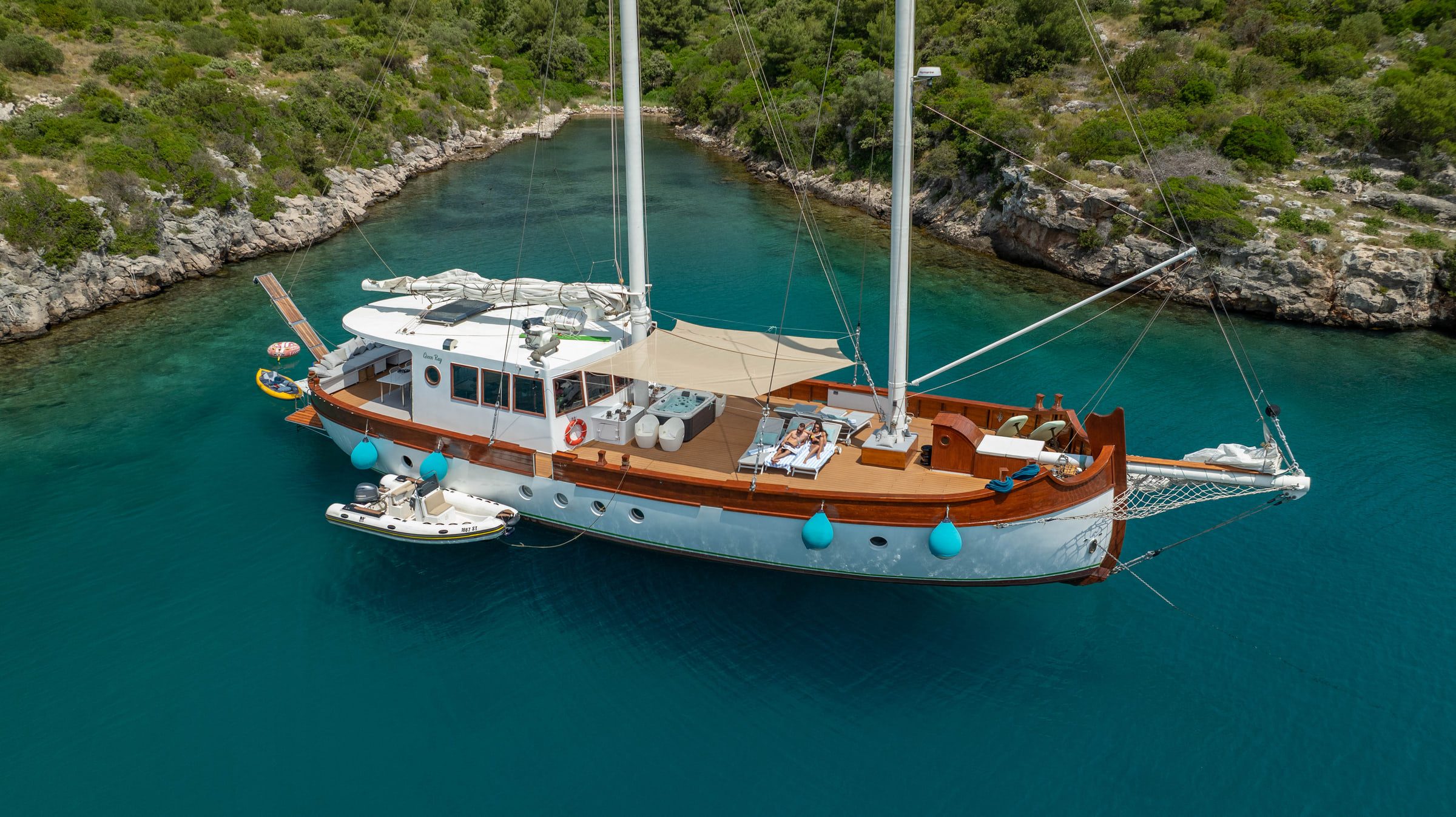 Seen from above, the Green Ray lies at anchor in a narrow turquoise cove, her tender alongside.