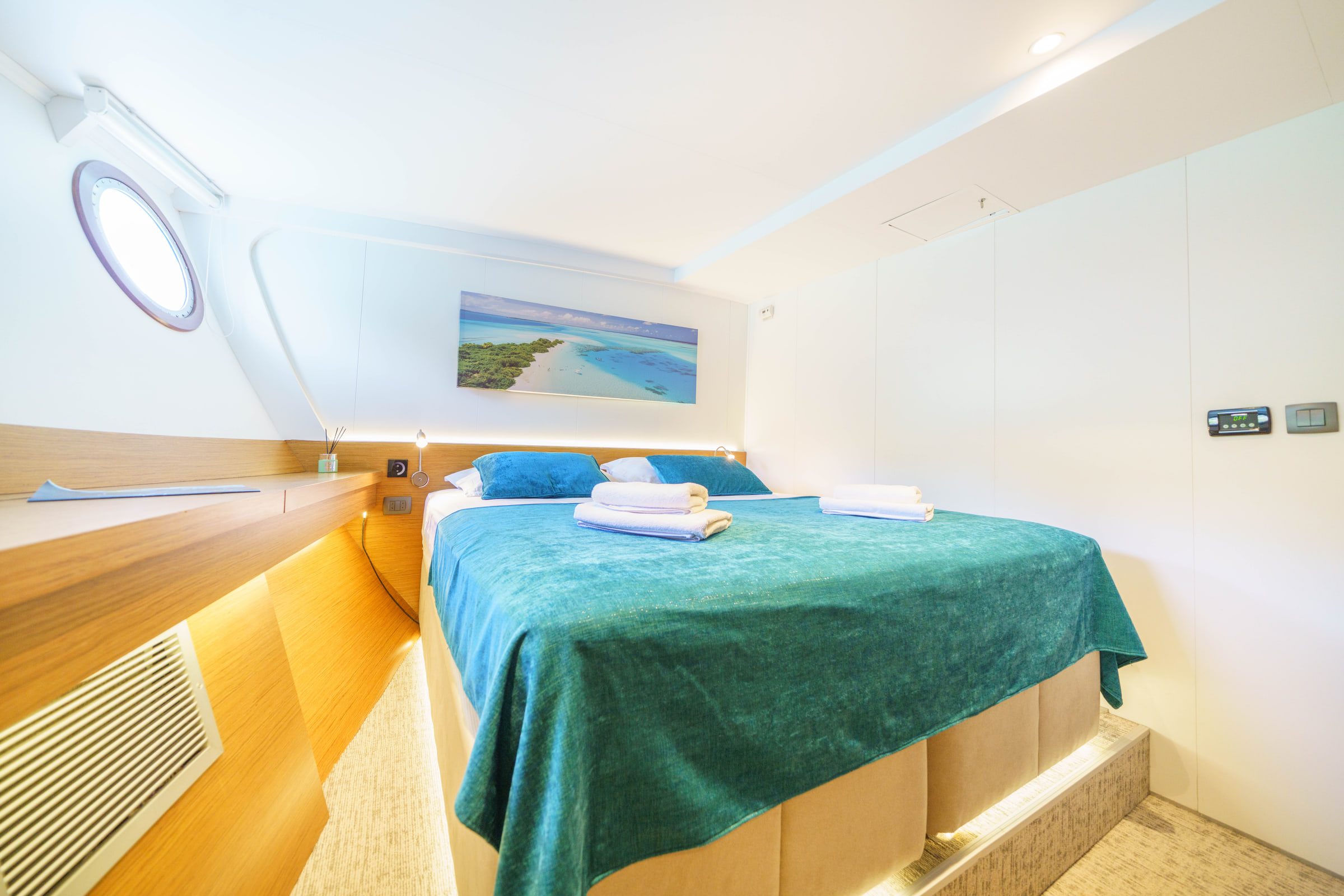 A double cabin with a teal bedspread, a porthole and a beach print above the bed.