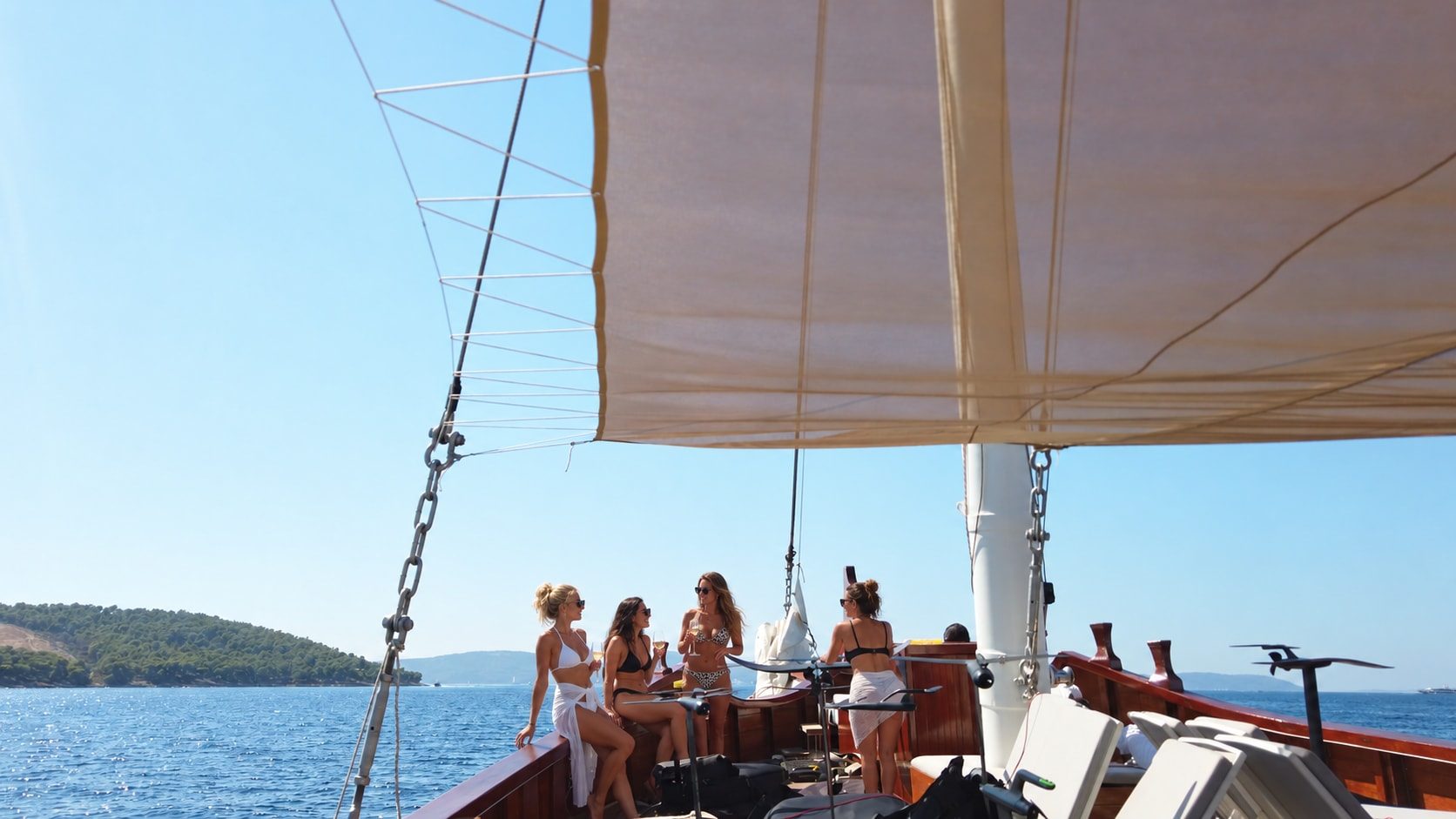 Guests in swimwear chat over drinks on the wooden foredeck under a white sun awning, a green island on the horizon.
