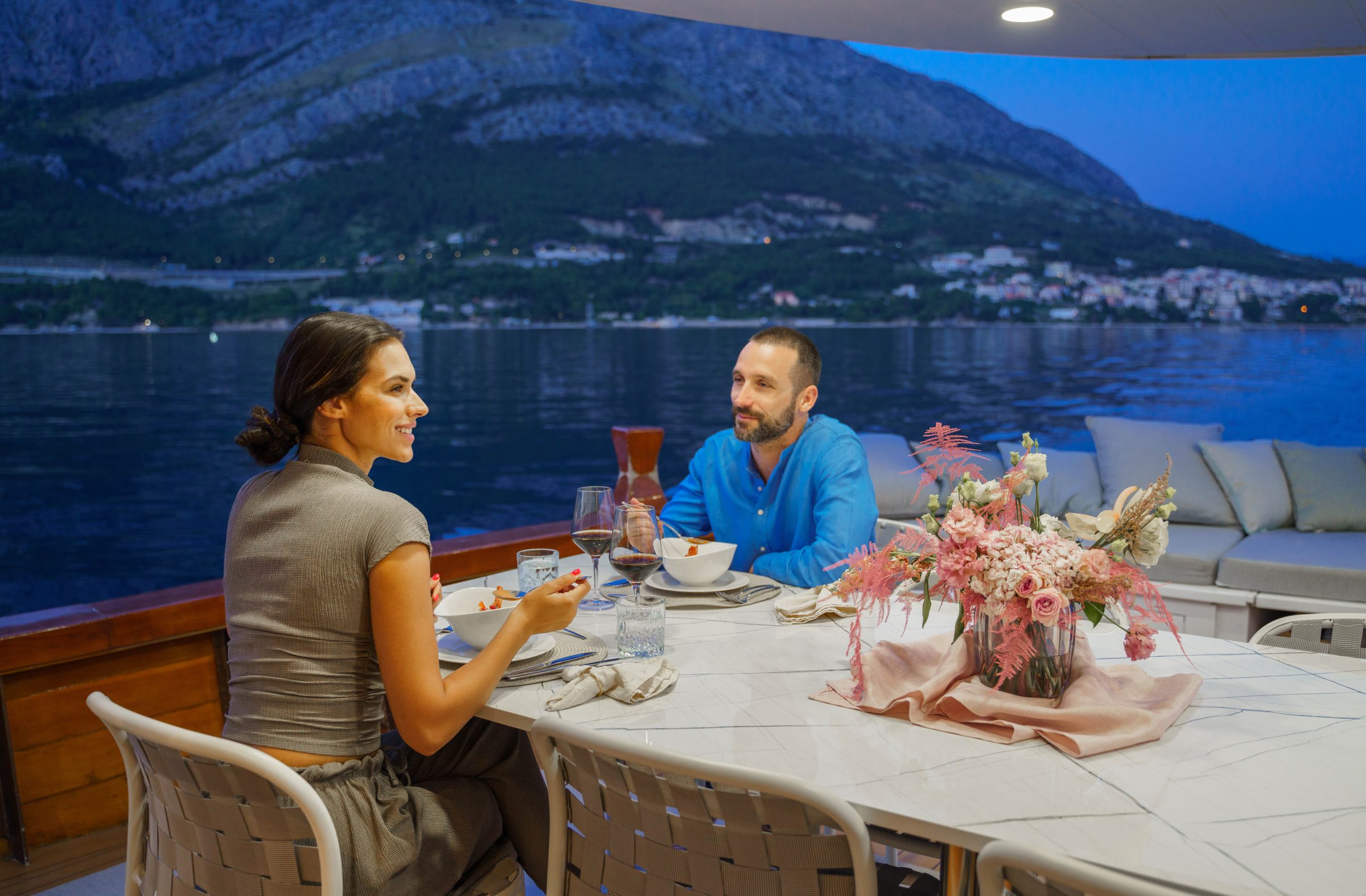 Two guests at dinner on deck at dusk, flowers on the table and the lights of a coastal town behind them.