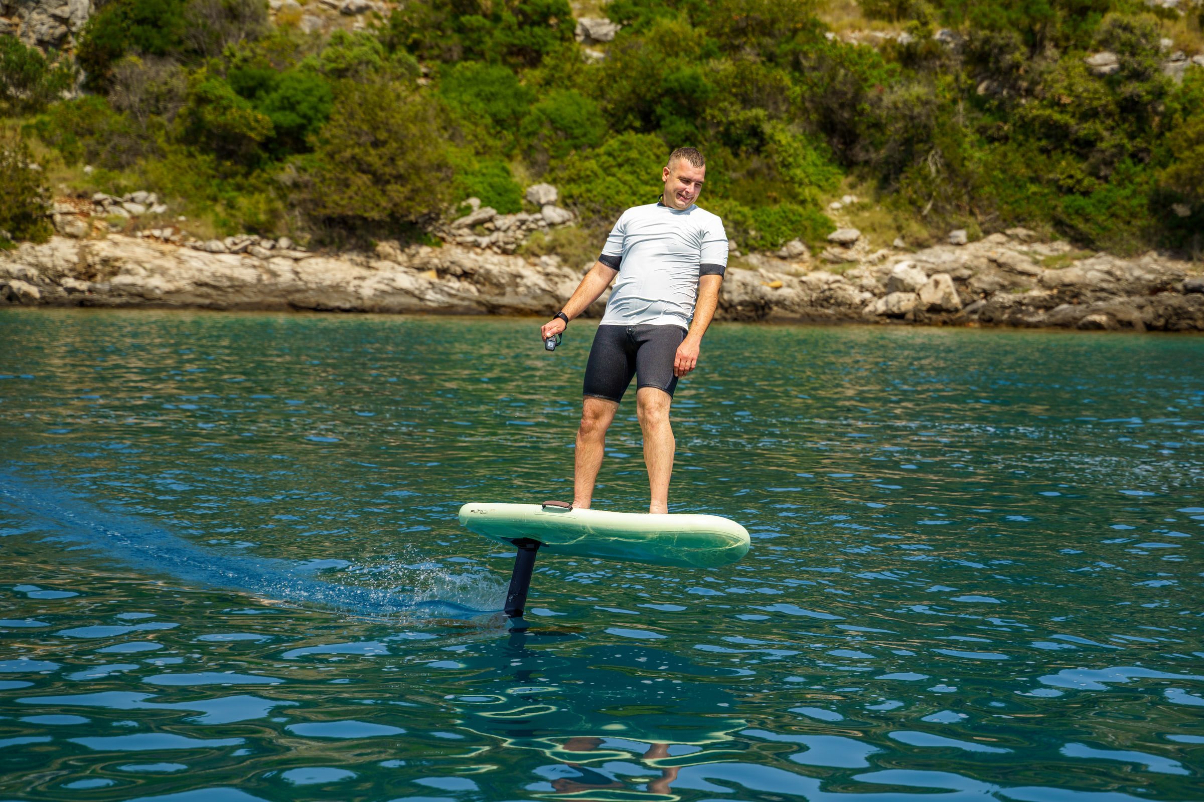 A rider stands relaxed on an eFoil, the board lifted clear above the blue-green water of a quiet bay.