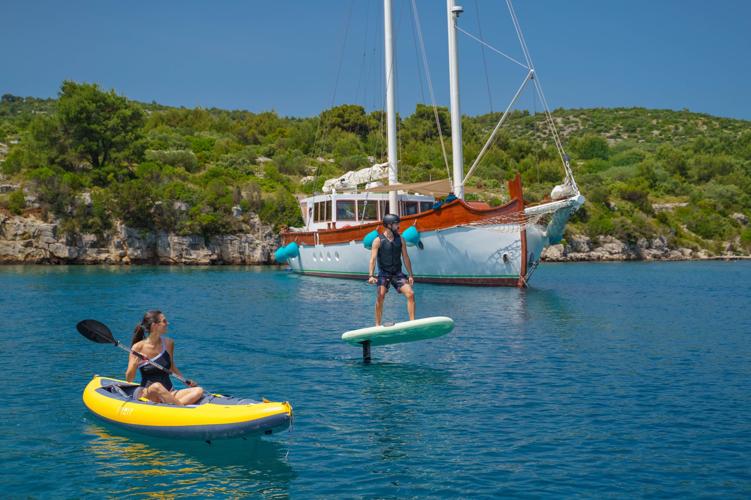 In a cove, an eFoil rider and a guest in a yellow kayak share the water in front of the anchored Green Ray.
