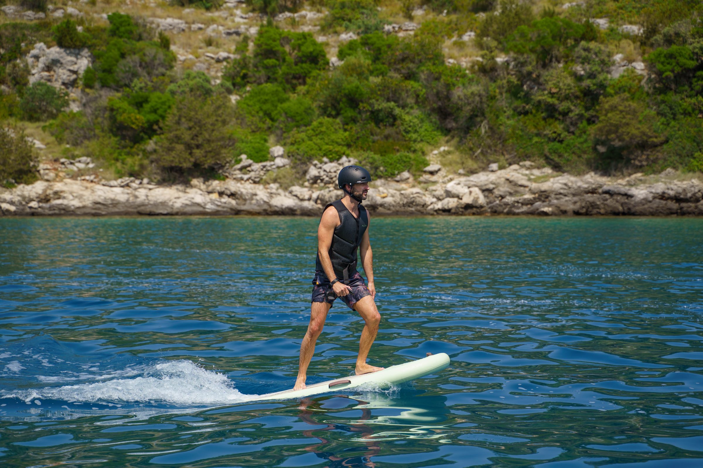 A rider in a helmet and life vest flies an eFoil low over clear water in a bay lined with pines and rocks.