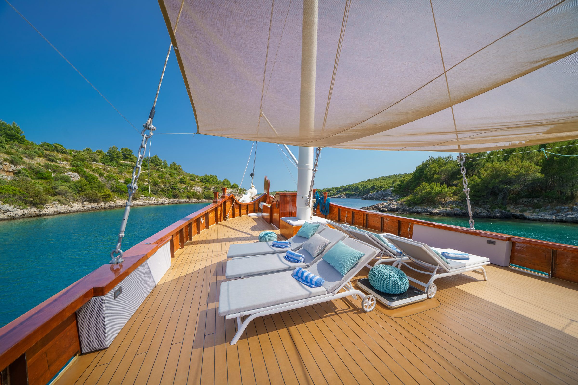 White sun loungers on the teak foredeck under a cream awning, a green shore just beyond the rail.