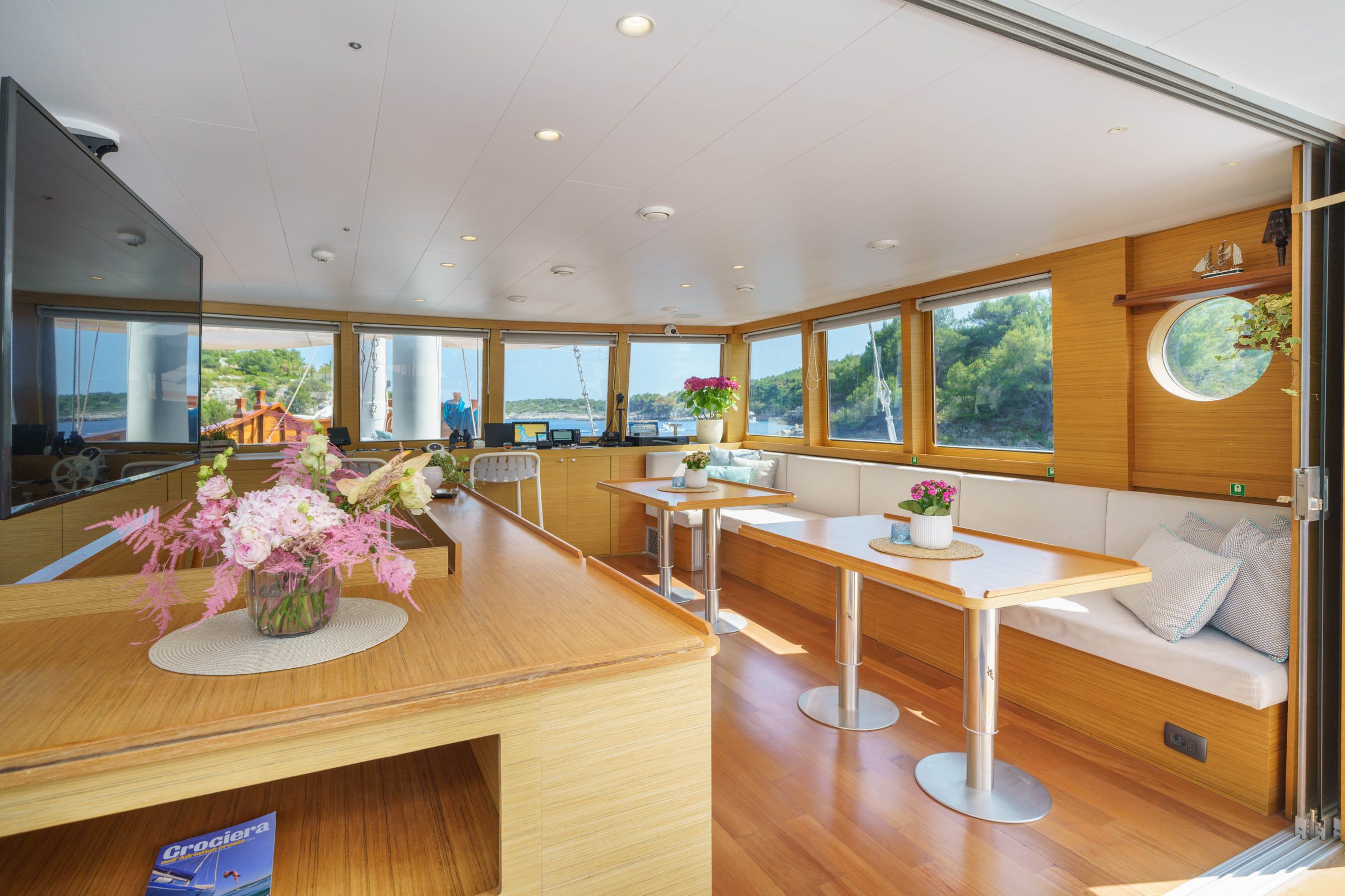 The bright salon with warm wood, round portholes, a long sofa and tables set with flowers.