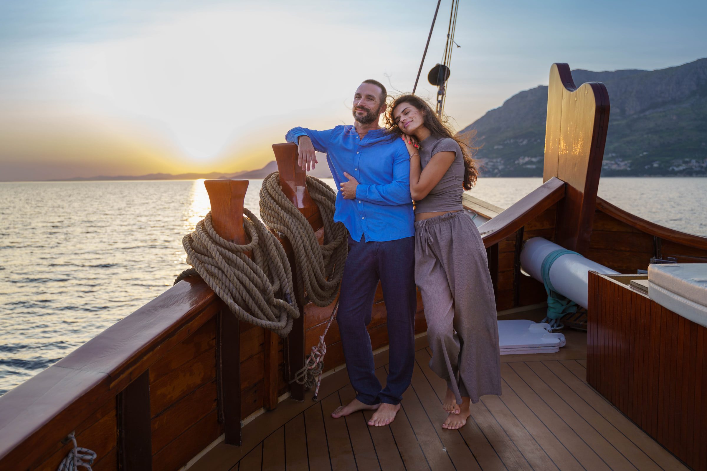 A couple leans on the bow rail at sunset, mooring ropes coiled beside them and mountains on the horizon.