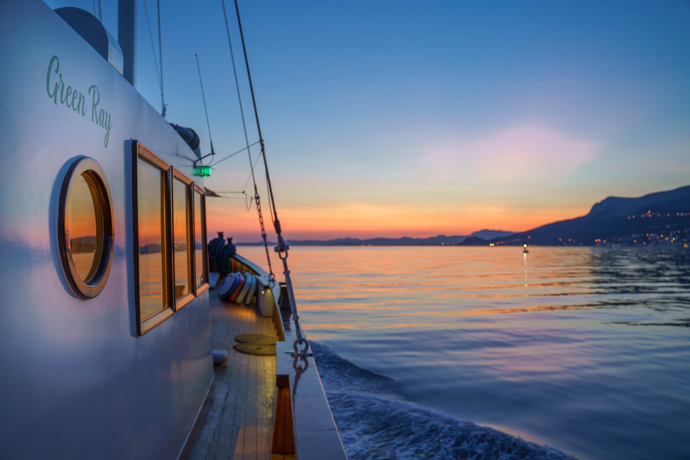 The Green Ray's white hull and round portholes at sunset, the coastline glowing on the horizon.