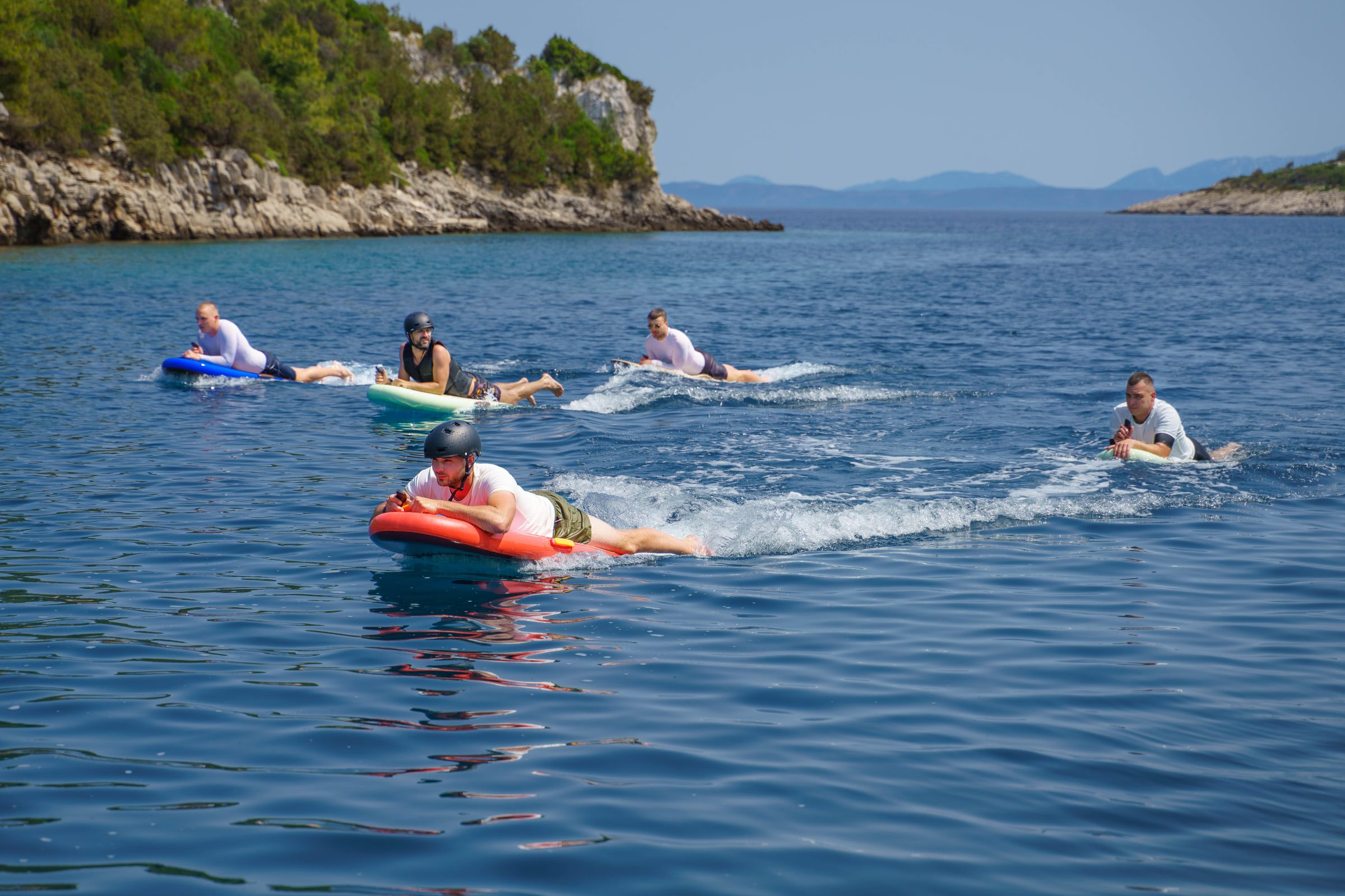 Guests ride inflatables and boards across open water near a rocky headland.